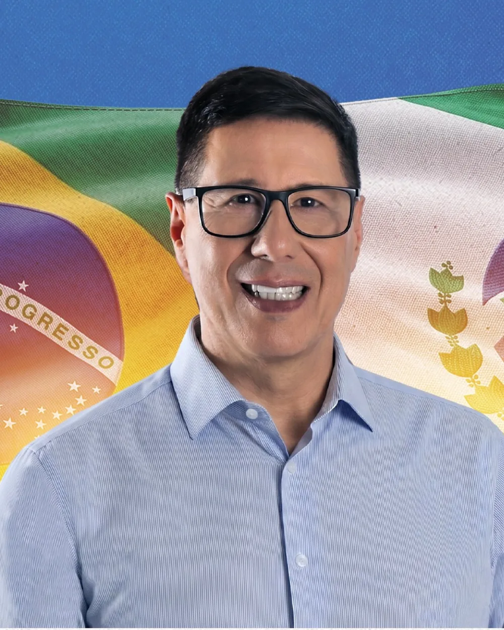 Retrato de Adão Litro sorrindo, de camisa azul clara, diante das bandeiras do Brasil e do Paraná